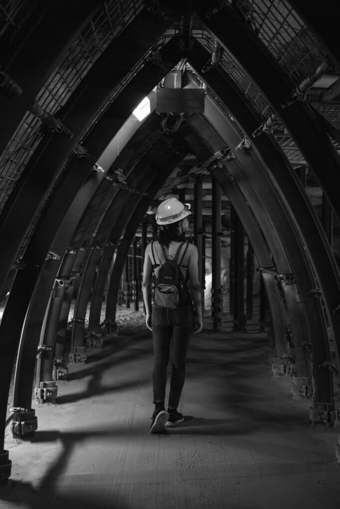 A woman in a hard hat and backpack walks through an industrial tunnel in grayscale.